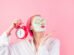 Young woman with cosmetic mask on face with cucumber slices to eyes. Beautiful woman with facial mask and alarm clock in hand. Beauty treatment. Facial mask. Spa therapy. Relax. Woman with facial mask
